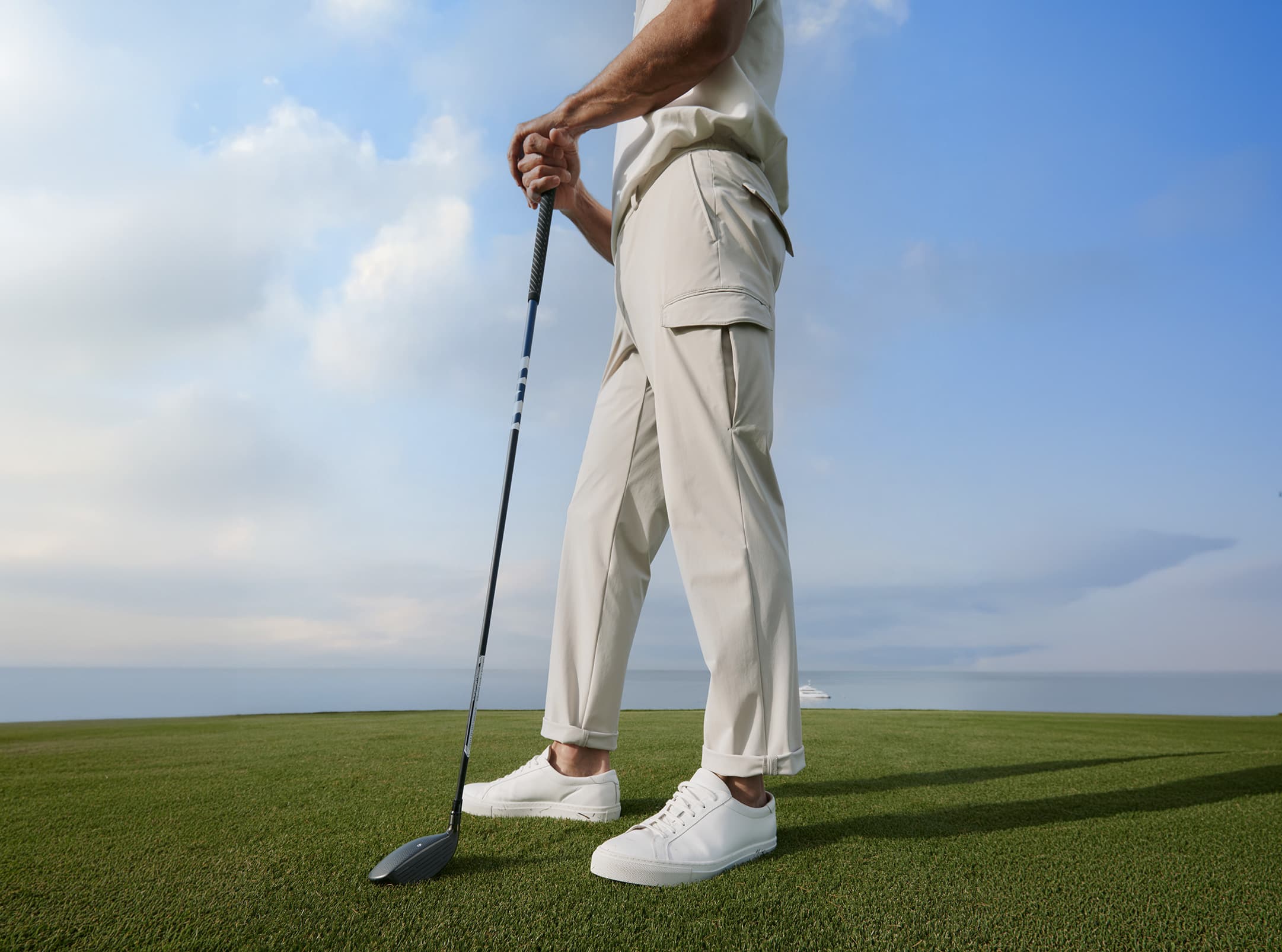 Nahaufnahme eines Mannes auf einem Golfplatz, der einen Golfschläger hält. Im Fokus steht die Hose „Pebble“ in hellem Farbton mit modernem, geradem Schnitt und seitlicher Cargotasche. Der leichte Stoff und die klare Silhouette werden durch die stehende Haltung und die ruhige Naturkulisse betont.