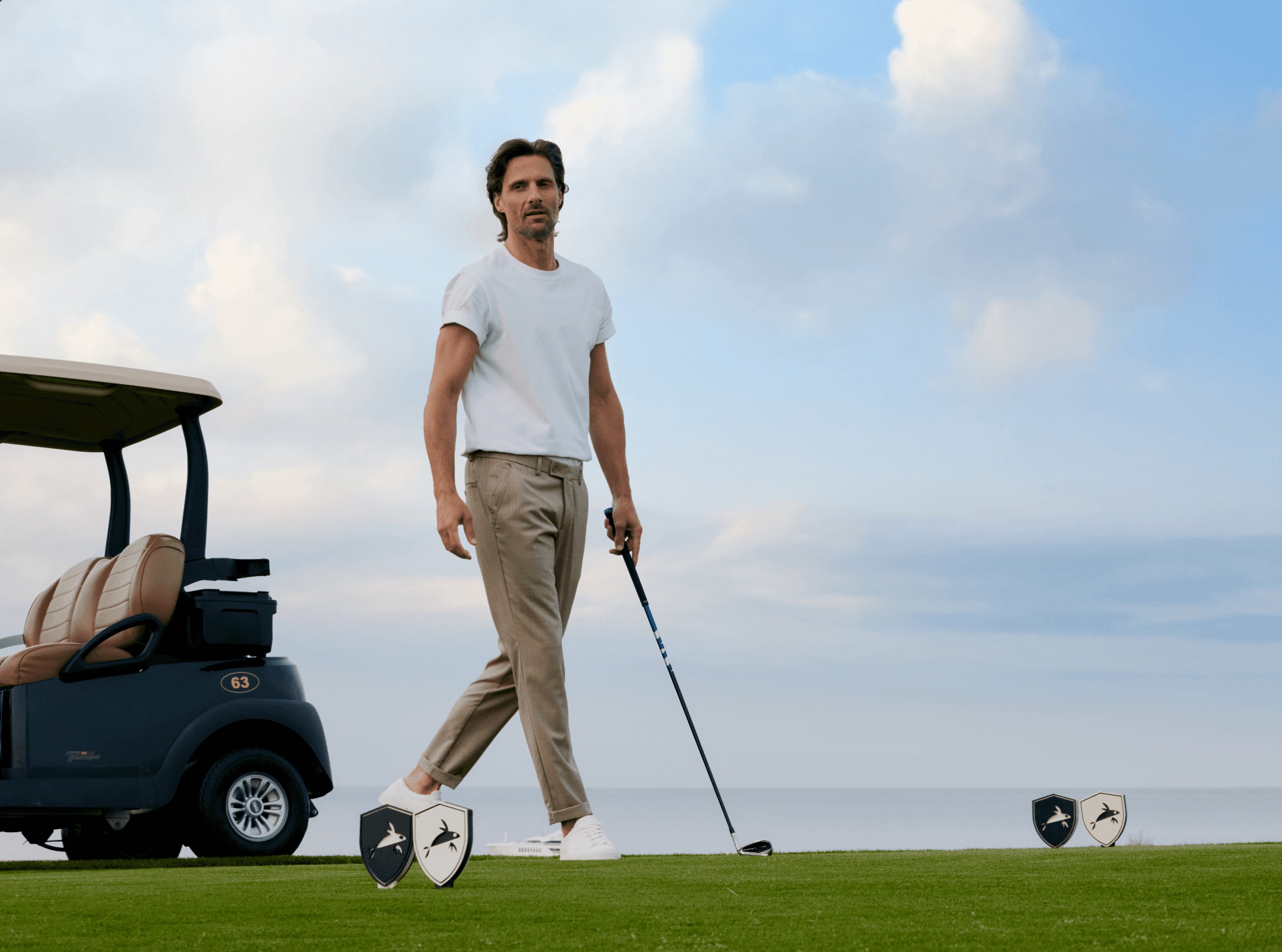 Ein Mann geht entspannt über einen Golfplatz und hält einen Golfschläger in der Hand. Er trägt die Hose „Turnberry“ in hellem Beige mit schlankem, modernem Schnitt, die einen eleganten und zugleich sportlichen Look vermittelt. Im Hintergrund steht ein Golfcart, während die offene Landschaft und der Himmel die ruhige, hochwertige Atmosphäre unterstreichen.