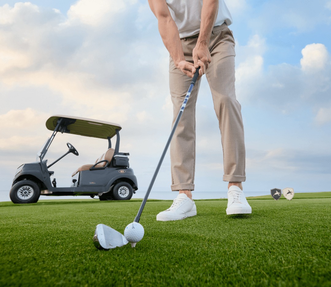 Ein Mann steht auf einem Golfplatz und bereitet sich auf einen Abschlag vor. Im Fokus steht die Hose „Turnberry“ in hellem Beige mit schlankem, modernem Schnitt, die Bewegungsfreiheit und einen gepflegten, sportlich-eleganten Look vermittelt. Im Hintergrund ist ein Golfcart zu sehen, das die Szene in einem gehobenen Freizeitkontext verortet.