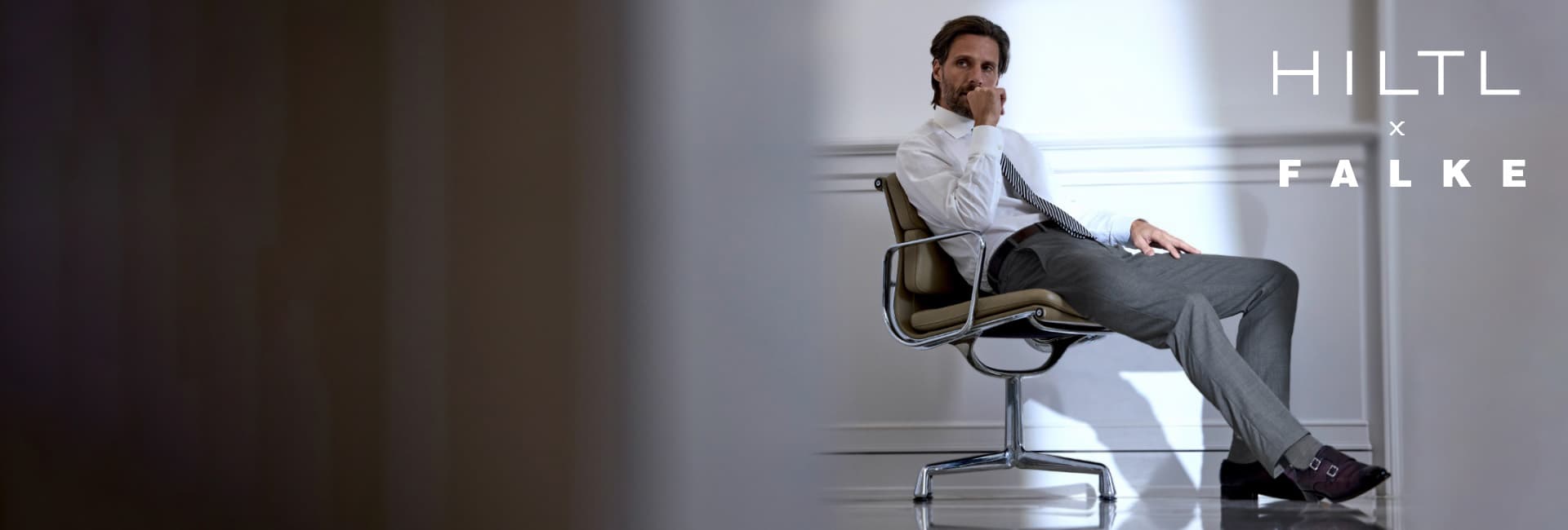 Ein Mann in Businesskleidung sitzt nachdenklich auf einem Designerstuhl in einem hellen, minimalistischen Raum. Er trägt ein weißes Hemd, eine Krawatte und graue Anzughose. Rechts im Bild stehen die Markennamen Hiltl x Falke.