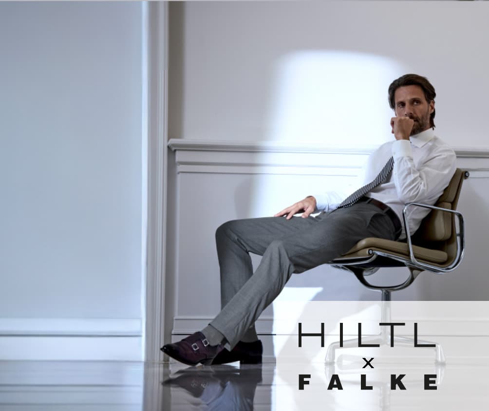 Ein Mann in Businesskleidung sitzt nachdenklich auf einem Designerstuhl in einem hellen, minimalistischen Raum. Er trägt ein weißes Hemd, eine Krawatte und graue Anzughose. Rechts im Bild stehen die Markennamen Hiltl x Falke.