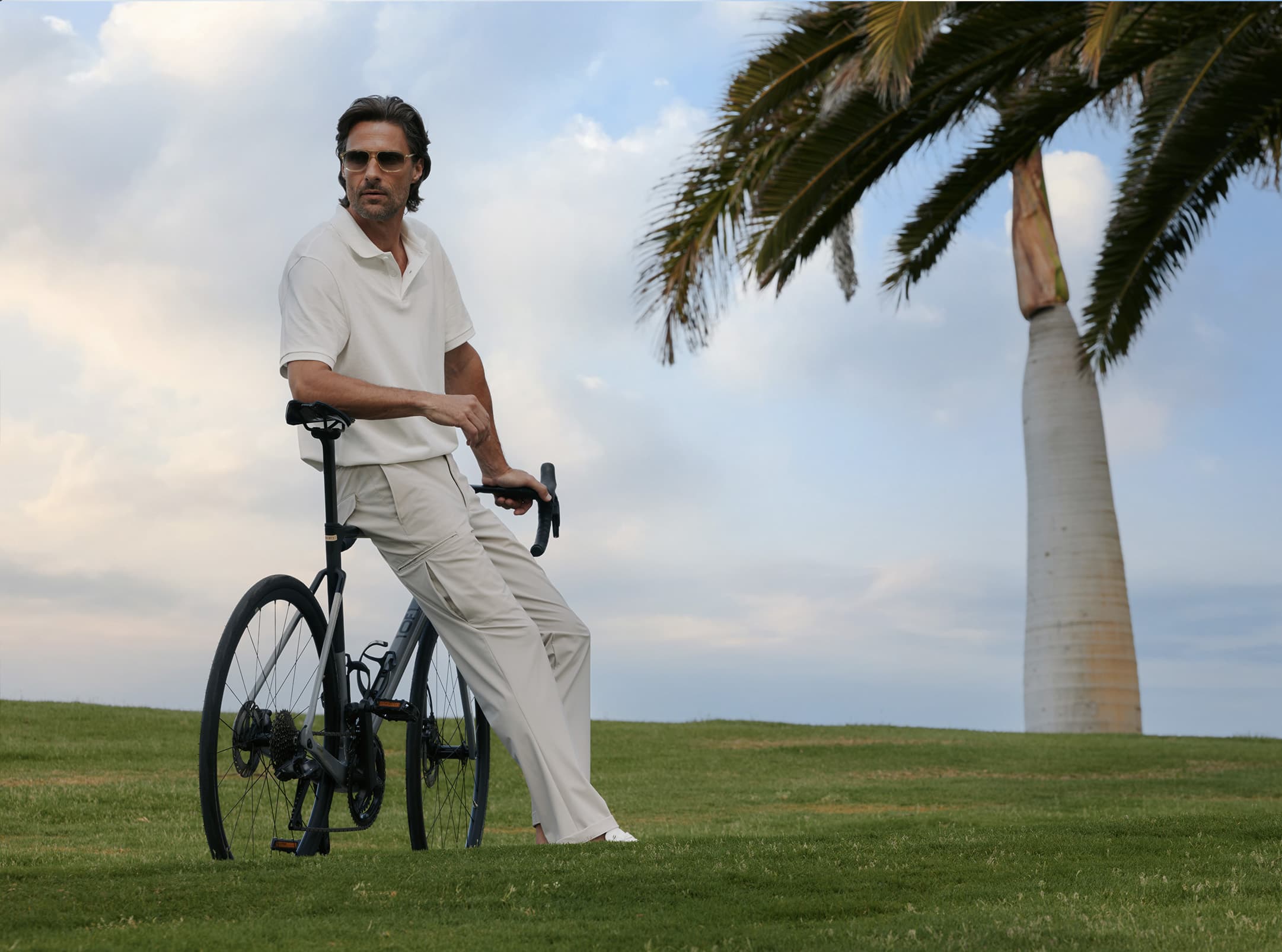 Eleganter Mann in einem monochromen, hellen Outfit lehnt entspannt an einem Fahrrad auf einer grünen Wiese, während im Hintergrund eine Palme im Wind steht. Er trägt die Hose „Pebble“ in einem fließenden, geradlinigen Schnitt, der einen luftigen und zugleich stilvollen Look vermittelt.